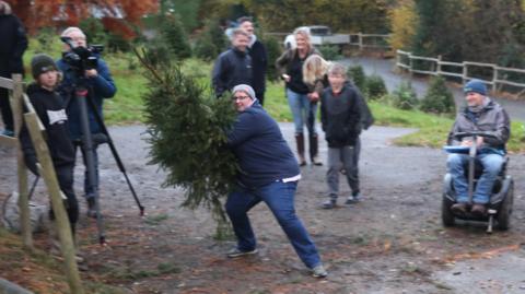 a man wearing blue trousers and blue jeans with a grey hat. he is leaning back, getting ready to throw a green Christmas tree. He is outside and surrounded by eight people watching