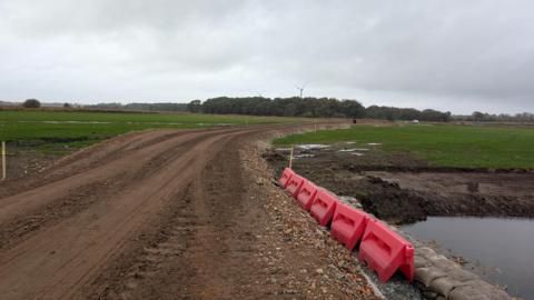 A dirt road has been created with fields either side and trees and turbines seen in the distance. Plastic red posts are at the side of the track with sandbags by water. The sky is cloudy.
