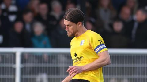 Charlie Raglan, wearing a yellow Barrow away kit, passes the ball with his right foot in the FA Cup tie at Spennymoor