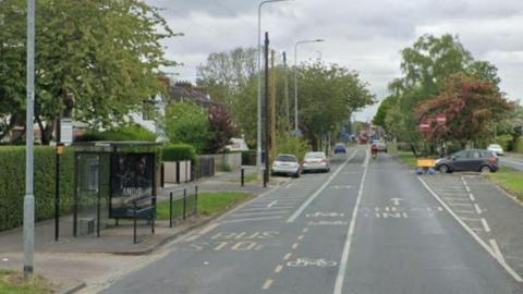 A bus shelter next to a tree-lined road with residential housing on the left of the picture