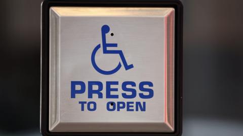 Close-up of a stainless-steel accessibility door button displaying a blue wheelchair symbol above the words “PRESS TO OPEN.” The square button is mounted on a post, with a blurred indoor background.