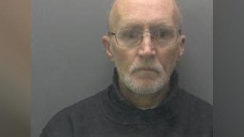 Peter Swan's custody photo. He is wearing a black or navy turtle neck jumper. He is bald and has glasses on.