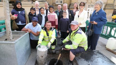 Two men wearing hi-vis yellow jackets hold a spade and a large metal cylinder as they crouch down next to a hole in concrete. A group of children and adults stand behind them looking at the camera. 