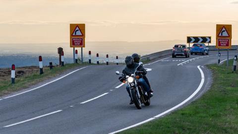 A motorbike and passenger in black leather ride towards the camera on a country lane. In the background are road signs and two cars travelling in the opposite direction