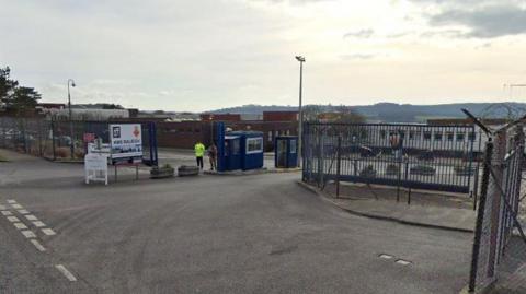 Two Royal Navy personnel stand guard at the main entrance to HMS Raleigh in Torpoint. There is a large sign with HMS Raleigh written on it and the gates and security huts at the entrance are navy blue.