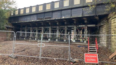 Metal fencing and red traffic sign with the words "Footway closed" under a bridge over the road