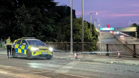 A police car is parked by police tape which is across a damaged barrier over a motorway in the evening