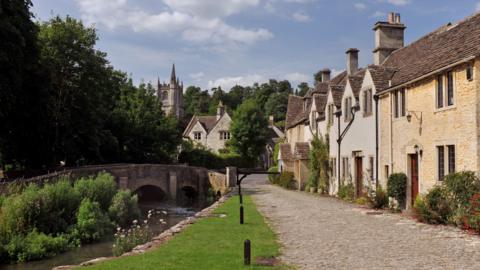 A view of Castle Combe on a sunny day, showing quaint cottages to the right, a bridge and shallow river and a church steeple in the background with trees