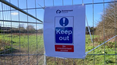 A laminated A4 sign reading: Somerset Council, keep out, no unauthorised access. It is cable-tied to a metal fence in front of a field.