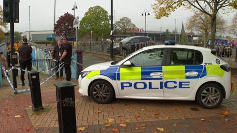 Several police officers are standing beside a police car, which is parked on a pavement. There are black bollards in the foreground and police tape. There are trees and a Sainsbury's supermarket in the background.