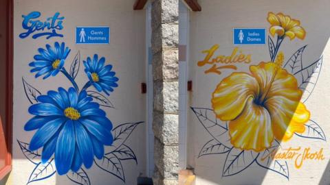 Close-up of two painted walls by public toilets side by side. The left door is labelled “Gents Hommes” and decorated with large blue flowers; the right door is labelled “Ladies Dames” and decorated with large yellow flowers. The artwork is painted directly onto the wall.