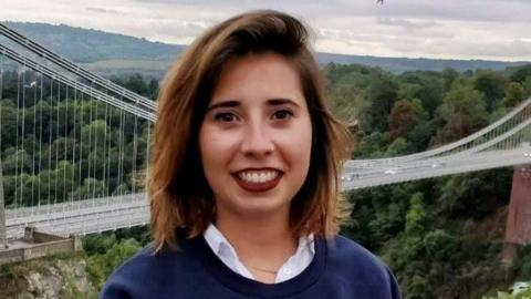 Marta Elena Vento smiles at the camera. Clifton Suspension Bridge is in the background. She has shoulder-length brown hair and wears a white shirt, blue top and make-up.