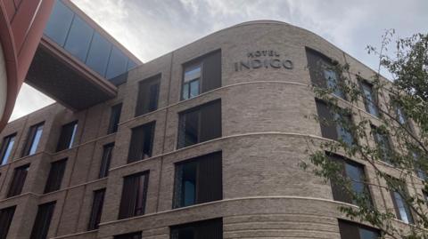 A large grey brick building with several storeys. There are modern windows and curved walls, with a red metal bridge leading to the top floor from another building. The sky above is grey.