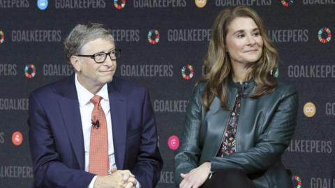 Bill and Melinda Gates seen at an event. They are both looking away but are seated on stools on a stage. 