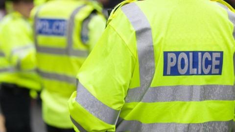 Three police officers standing outside with their backs to the camera. They are all wearing yellow fluorescent jackets which have silver stripes on the back and arms and writing which says POLICE on the back. 