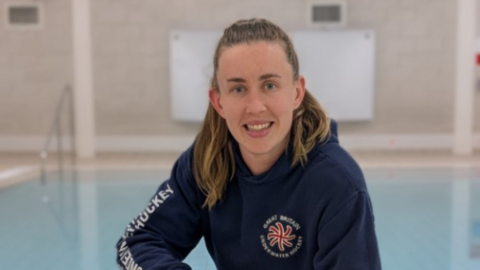 A woman wearing a blue hoodie and smiling at the camera. She is stood in front of a swimming pool.