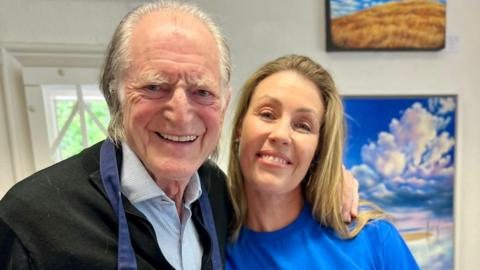 Two people smile in a room with white walls and a landscape painting of a boat hung behind them. On the left is an elderly man with white hair and round wire-framed glasses, wearing a black jumper and blue shirt, with a painting apron on top. To his right is a woman with long blonde hair wearing a blue t-shirt.