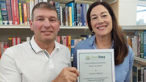 A man with short brown hair with a small patch of grey at the front and wearing a white short-sleeved polo shirt with black trim around the collar holds a certificate from Read Easy as he smiles into the camera. A woman next to him, with long dark brown hair, pearl earrings and a blue and white-striped shirt, grins broadly in to the camera