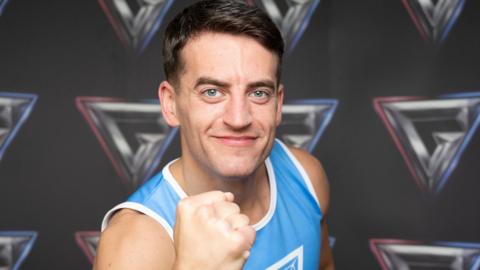A person wearing a blue and white athletic vest stands with arms crossed in front of a backdrop displaying multiple geometric logos.
