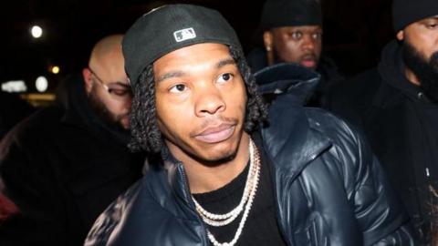 Lil Baby pictured in January 2025 at a launch event for his new album. He wears a black cap twisted back and a black puffer jacket over a black T-shirt and jewelled necklaces. 