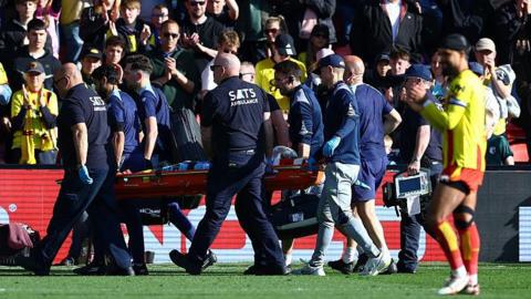 Footballer Conor Coady being carried off a football pitch on a stretcher by a team of male medics wearing dark colours while a player in a yellow shirt with red trim, red shorts, white socks and white boots is visible in the foreground and fans applauding are visible in the background