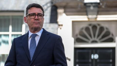 Andy Burnham wearing a dark suit, white shirt and blue tie. He has dark hair and glasses. Burnham is standing outside of 10 Downing Street.