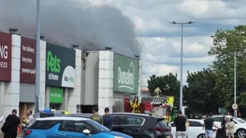 Retail park with smoke billowing from a Dunelm store, flowing over the roofs of neighbouring shops. There is a fire engine by the store entrance and cars parked in the car park. People are standing around watching the fire.