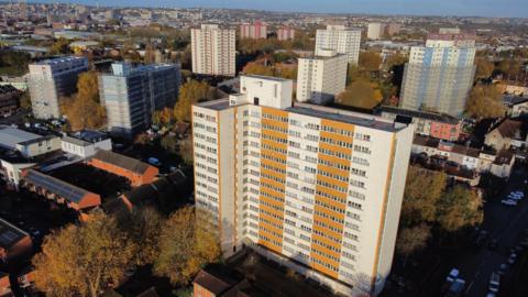An aerial shot of Barton House in Bristol. It is a large tower block with white and orange panels and lots of windows on it. Other towerblocks and residential properties can be seen in the distance and around Barton House.