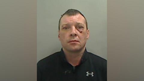 Mugshot of Carl Wild. The image is pixilated but he has several cuts to his face and a black eye. He has short brown hair and is wearing a black t-shirt with the Under Armour logo .