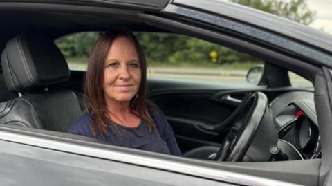Caroline O'Brien sitting in her car looking directly at the camera. She is wearing a blue top. She is visible from the shoulders up and is sitting in the driver's seat with the steering wheel in front of her. There is a road and foliage in the background.