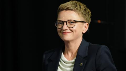 Bev Craig, wearing glasses and a black jacket over a white top, smiles into the camera
