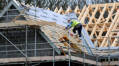 A man on a the roof of house being built. He is in a hi-vis vest and working on building the roof. There is a timber frame behind him and scaffolding beneath him.