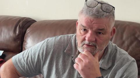 Neville Bradford (grey hair, beard, glasses on head) sits with his chin on his hand looking at camera. Wearing grey polo top.