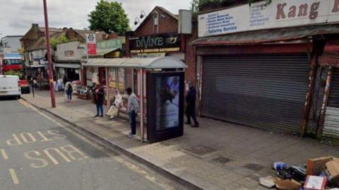 A generic shot of Soho Road, featuring people waiting in a bus shelter by the side of a road. Metal shutters are down on premises to the right and a pile of rubbish is on the pavement.