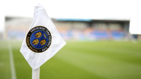 A corner flag with the Shrewsbury Town badge in the foreground of picture of Croud Meadow stadium 