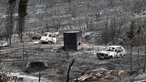 A scene of devastation after a wildfire swept through an area. everything is covered in ash and all vegetation is a black cinder. In the centre of shot, two burnt out cars sit.