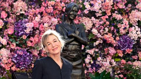 Renée Zellweger smiles while wearing a black dress and posing next to a statue of Bridget Jones against a backdrop of pink and purple flowers lining the wall, in Leicester Square in London on Monday.