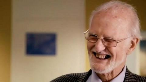 Keith has white hair and a white beard. He wears glasses and smiles broadly as he is interviewed.