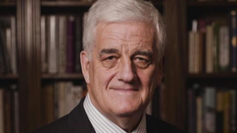 Man with short white hair looking at the camera, he is wearing a striped shirt and black jacket.