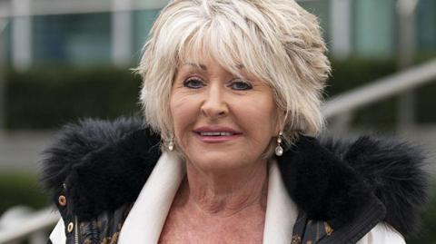 Former detective Maggie Oliver, who has short blonde hair, wears dangly earrings and a cream jacket with black furry hood. She is standing outside a large building.