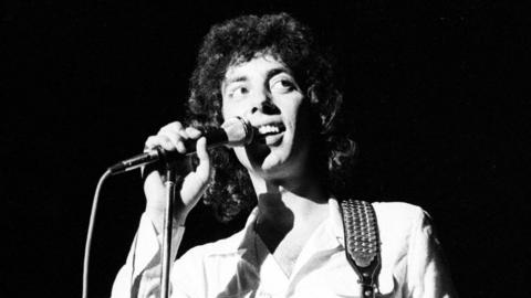 A black-and-white image of Graham Gouldman on stage in the 1970s, singing into a microphone. He has long curly hair and is wearing a shirt with a guitar strap over his shoulder.
