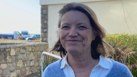 Jo Le Noury. She has shoulder length brown hair and is wearing a bluie jumper. 