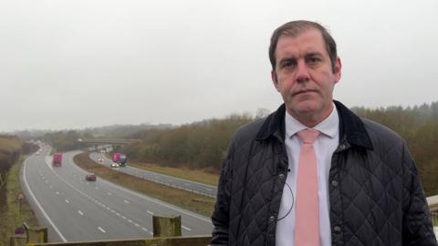 MP Lee Dillon stands on a bridge over the A34. He is wearing a black jacket with a white shirt and a pink tie. In the background both directions of the A34 are visible. 