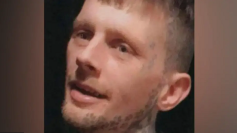 Pictured is Kyle Pugh. He has blonde hair and face tattoos along his jaws line and at the side of his temple.