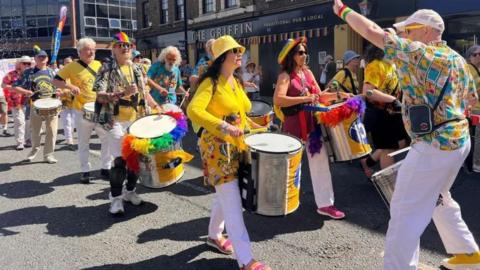 A Pride parade with men and women walking as they play drums, wearing yellow and other bright colours, including rainbow hats and feathers. 