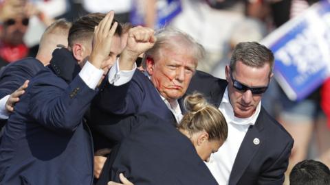 Donald Trump holds up his fist as blood is smeared across the left hand side of his face. He is flanked by secret service agents