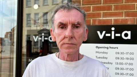 Mike Major looking directly into the camera. He has short grey hair and is wearing a white t-shirt. Behind him is a sign that says v-i-a and the charity opening hours. 