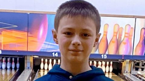 A young boy in a blue hoodie with short brown hair looks at the camera. Behind him is a bowling alley.