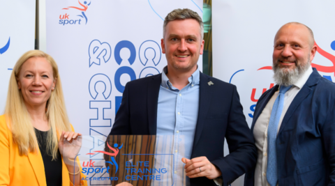 A woman with long blonde hair on the left is wearing a yellow jacket and standing next to a man in the middle with a shirt and dark blue jacket. A man with a grey beard on the right is wearing a shirt, tie and blue jacket.