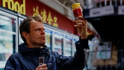 Dan Kelly holding up a jar of pickles and holding a dictaphone in his other hand. Some supermarket fridges are seen in the background.